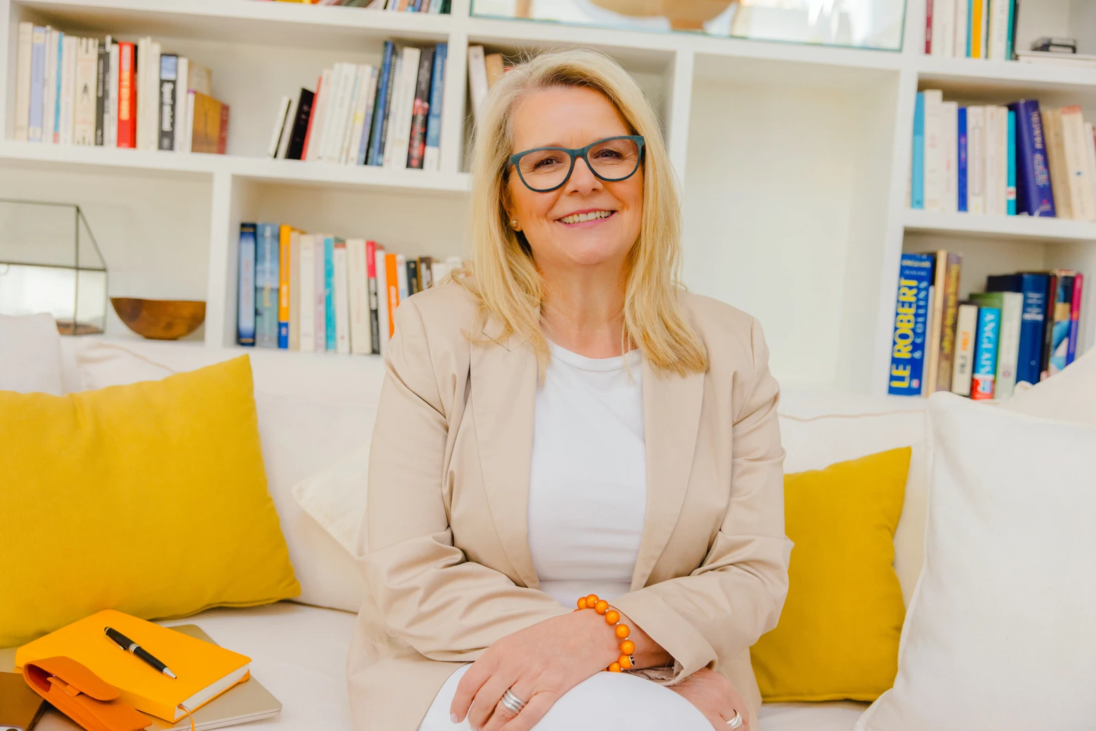 Unternehmerin mit dunkler Brille und beigem Blazer auf einem Sofa vor einem Bücherregal