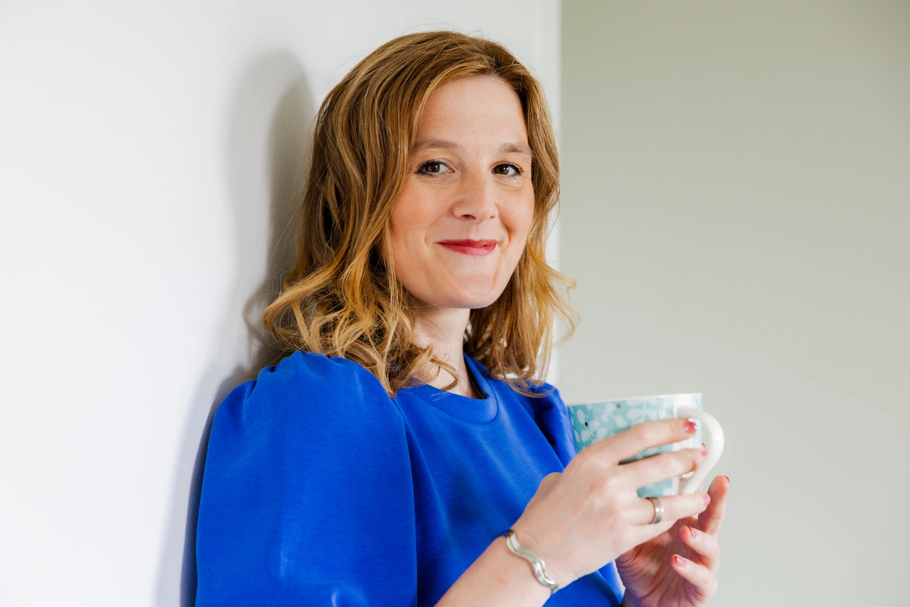 Businessportrait einer Unternehmerin im blauen Oberteil mit einer Tasse vor heller Wand