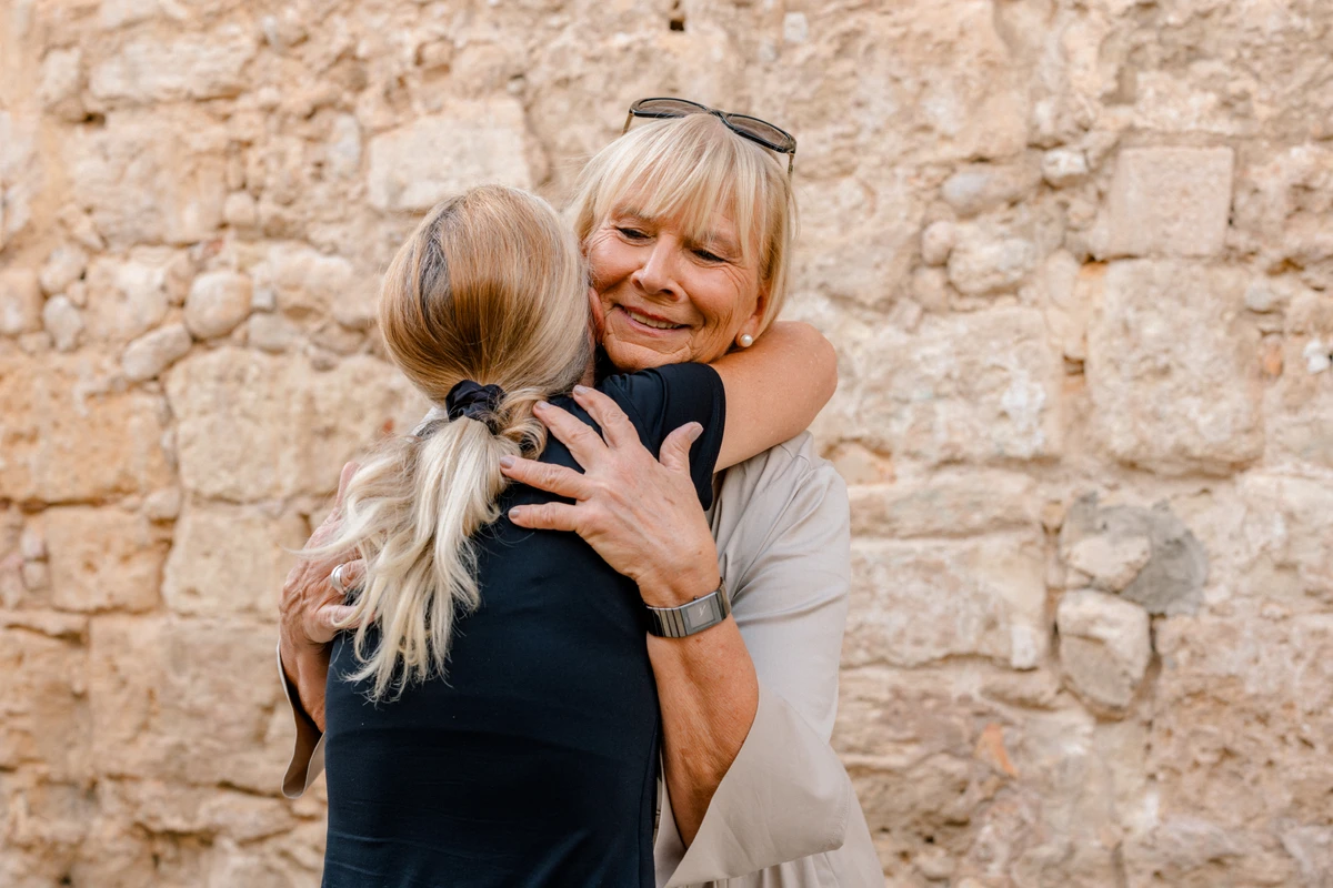 Unternehmerin umarmt eine Frau vor einer Steinwand. Fotografin: Manuela Engelking. Aufnahmeort: Mallorca.