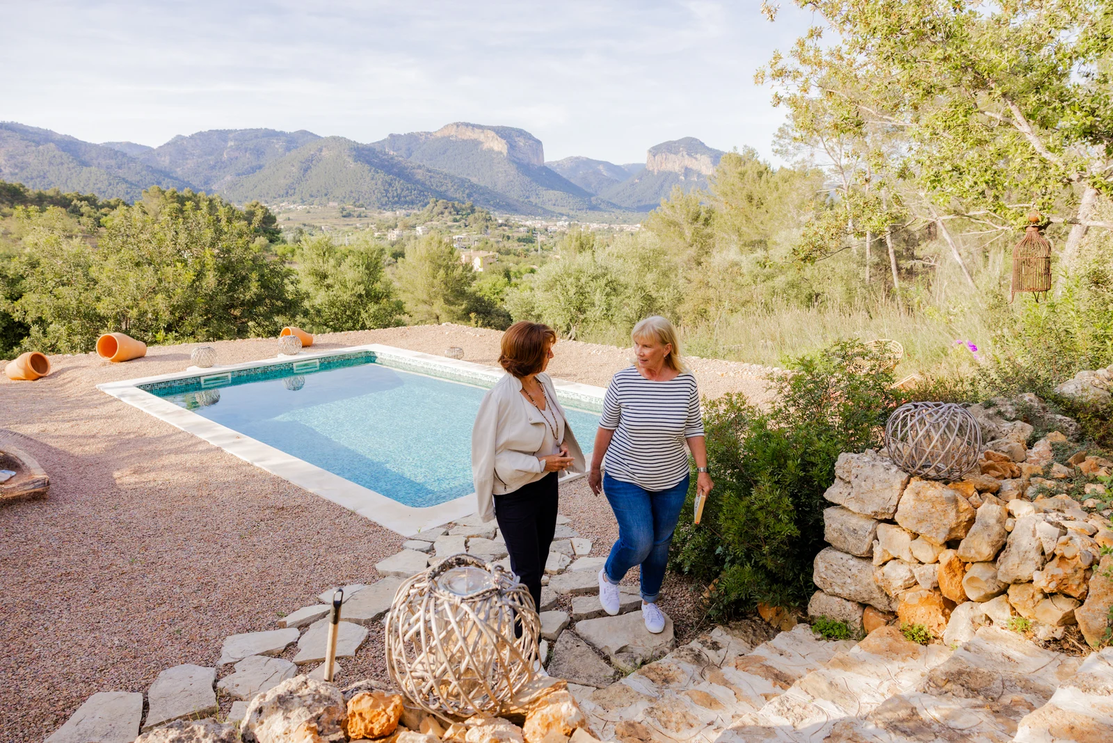 Zwei Frauen sprechen am Pool eines Hauses auf Mallorca; im Hintergrund sind die Berge zu sehen.