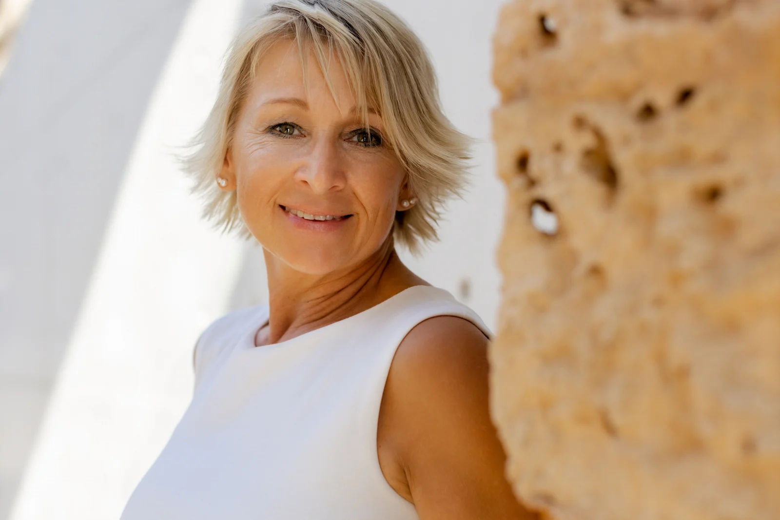 Portrait an heller Steinwand Unternehmensfotografie auf Mallorca: Blonde Frau im weißen Oberteil lächelt neben einer hellen Steinwand.