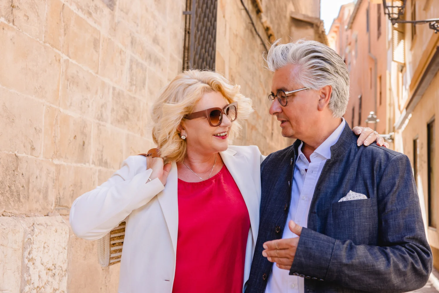 Gemeinsam in Mallorcas Gassen Unternehmensfotografie auf Mallorca: Frau im weißen Blazer und Mann im dunklen Sakko sprechen in einer Gasse.