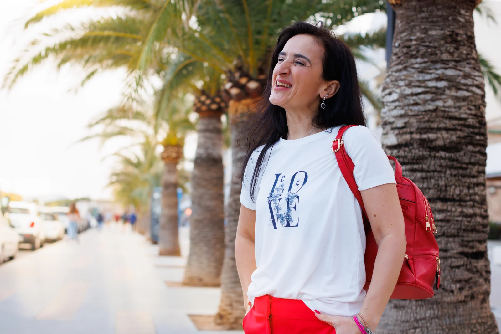 Unternehmensfotografie auf Mallorca: Frau mit weißem T-Shirt und rotem Rucksack lächelt unter Palmen.
