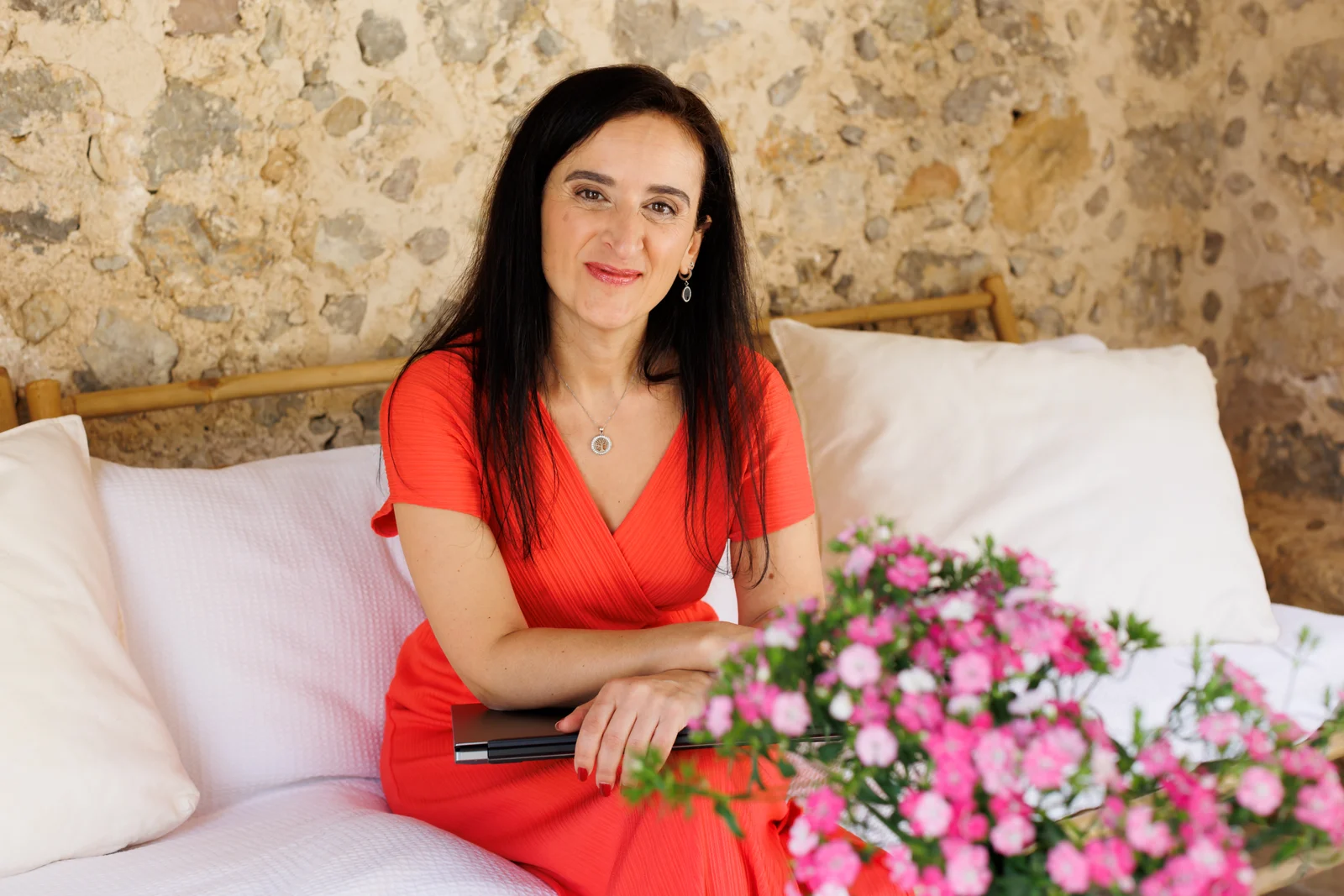 Unternehmensfotografie auf Mallorca: Frau im roten Kleid sitzt mit einem Notebook auf einem Sofa vor einer Steinwand.