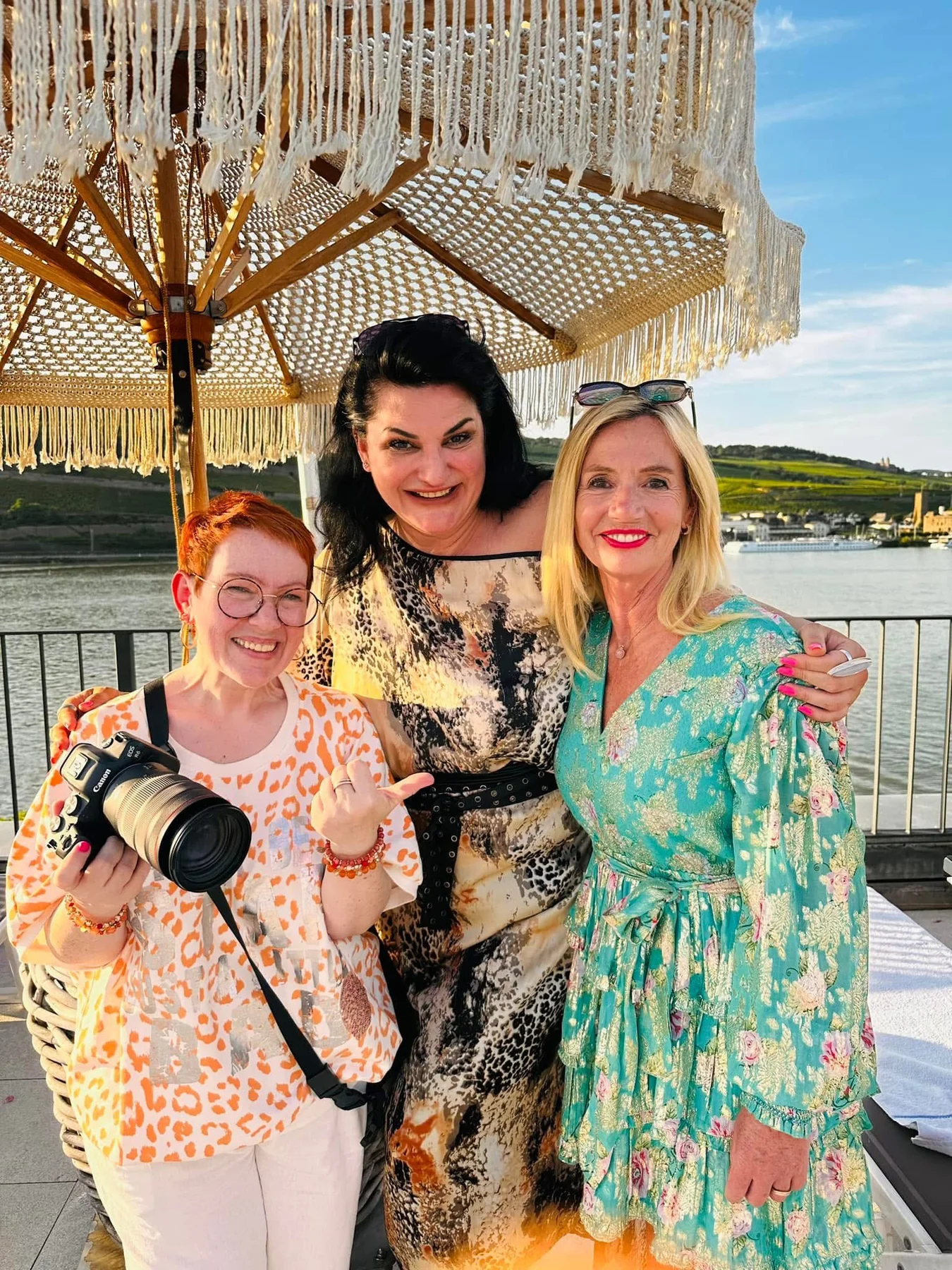 Manuela mit ihrer Kamera und zwei Frauen unter einem Sonnenschirm am Wasser
